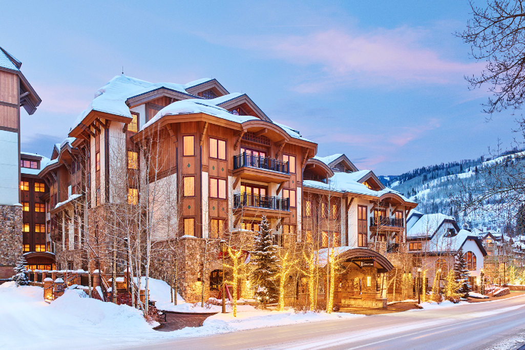The Sebastian Vail vacation rental seen from the outside in the early evening, showing off its stylish wooden finishings, fairy-lit trees at its entrance and snowy mountains in the background.