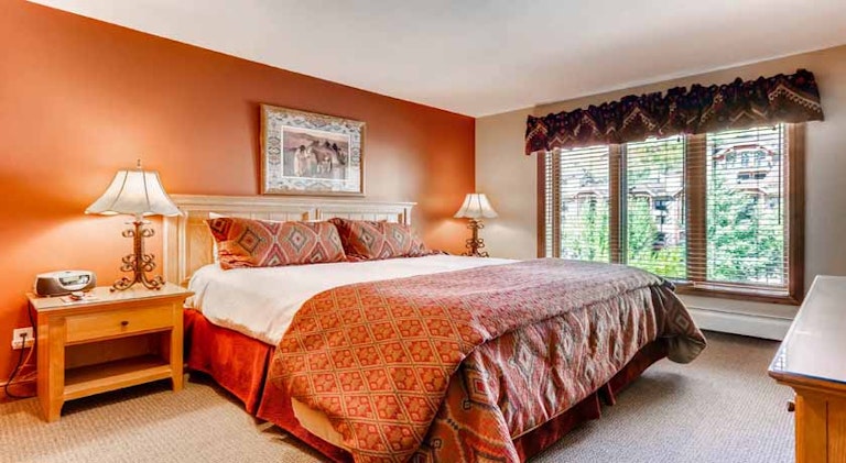 Vail condo rental option bedroom, with large bed, warm, orange color scheme and windows looking out onto greenery. 