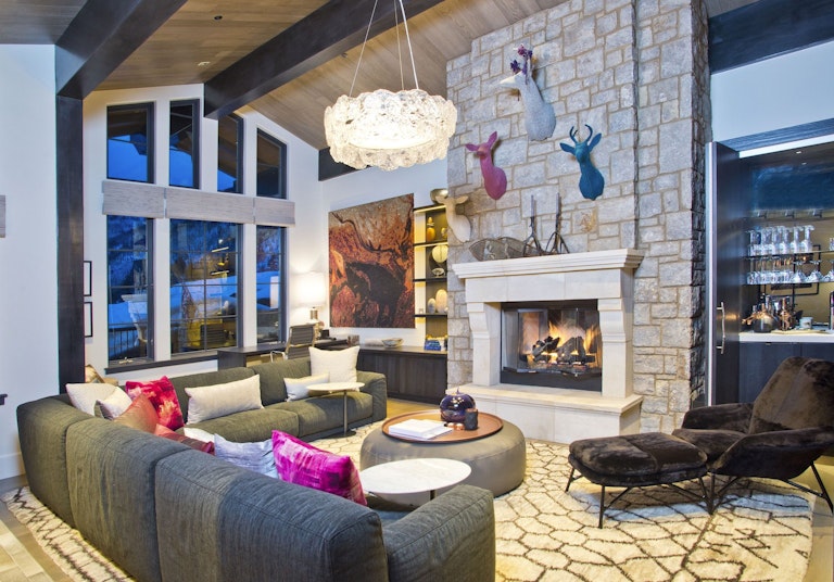 Vail Chalet Vail home rental's large lounge area with couches, lit fireplace, chandelier, large windows and a bar area visible in the background.
