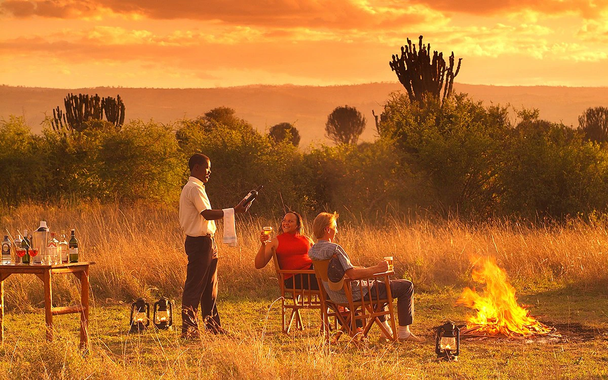 Two guests enjoy sundowners by an open fire as the sun sets around them