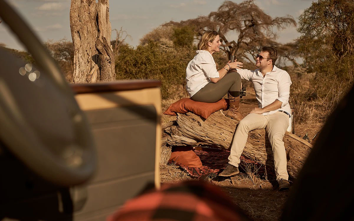 A pair of guests on a safari vacation take a break from their game drive to enjoy a drink sat atop a fallen tree branch