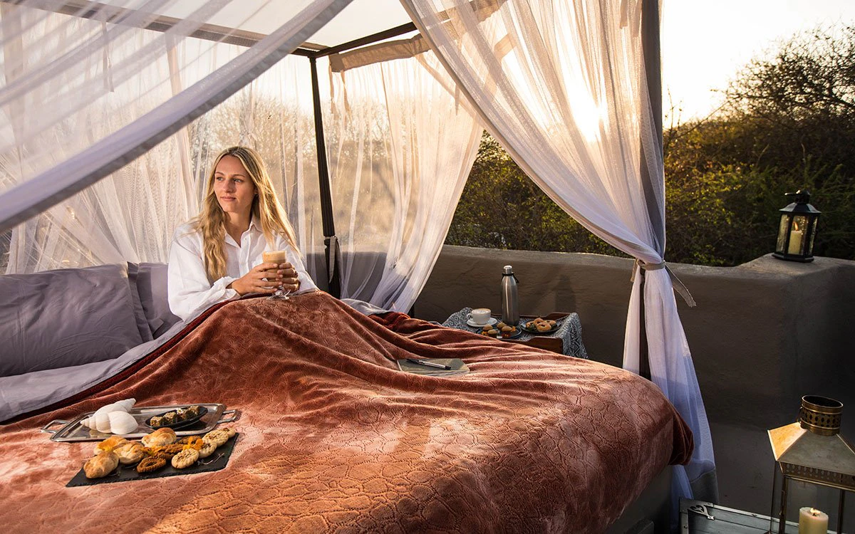 A lady relaxes in bed and enjoys a delicious breakfast, with the African landscape in the background