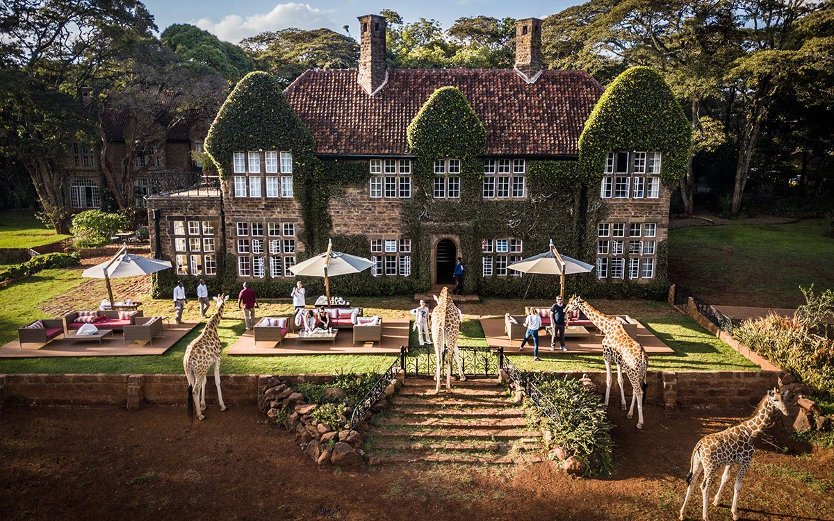 A front view of the exterior of Giraffe Manor with four giraffes walking in the foreground