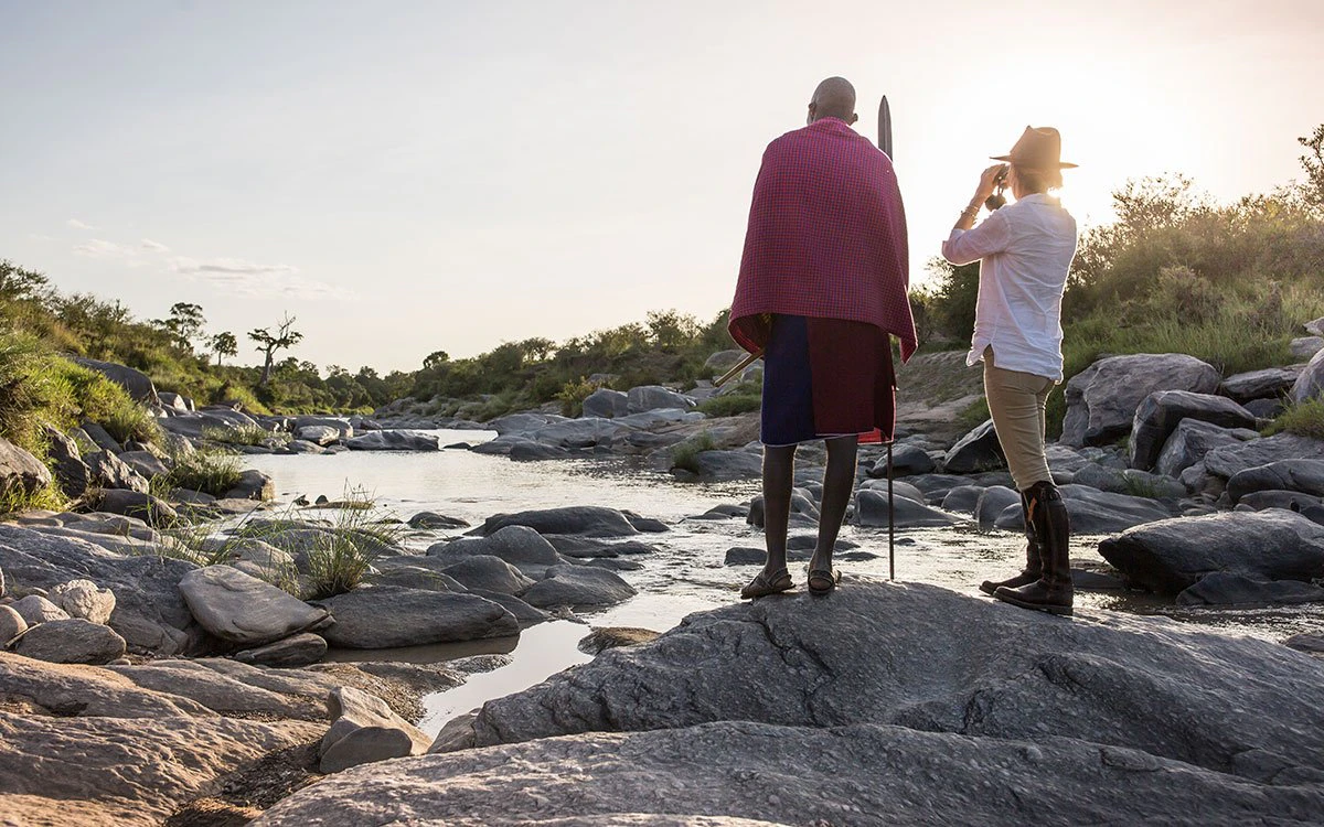 A guest looks down the Talek river using binoculars under the watch of a Maasai guide