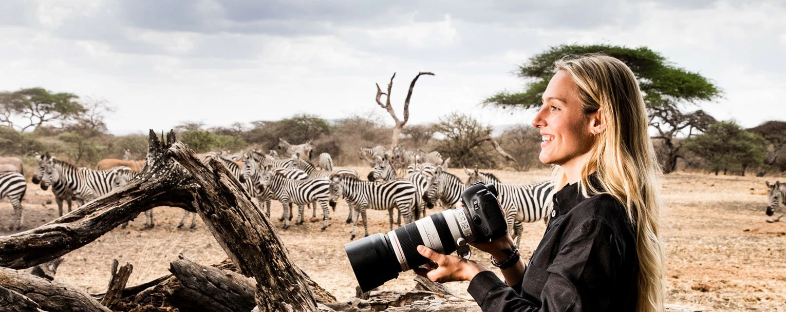 A woman on safari with her high powered camera with a herd of zebra in the background
