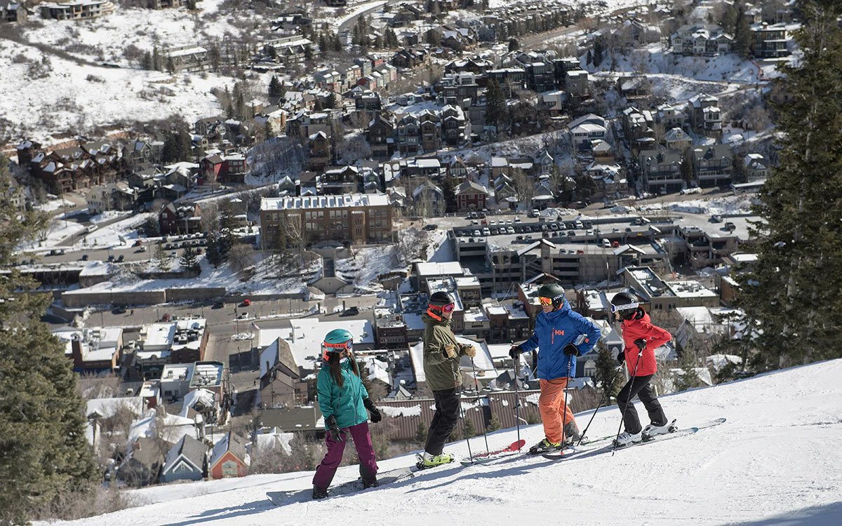 3 skiers and a snowboarder begin their descent down the mountain, with downtown Park City in the background.