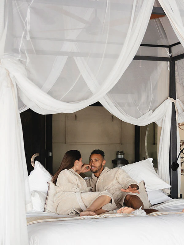 Couple relaxing in a luxury safari lodge bed in spa robes, enjoying a romantic moment during their African honeymoon retreat.