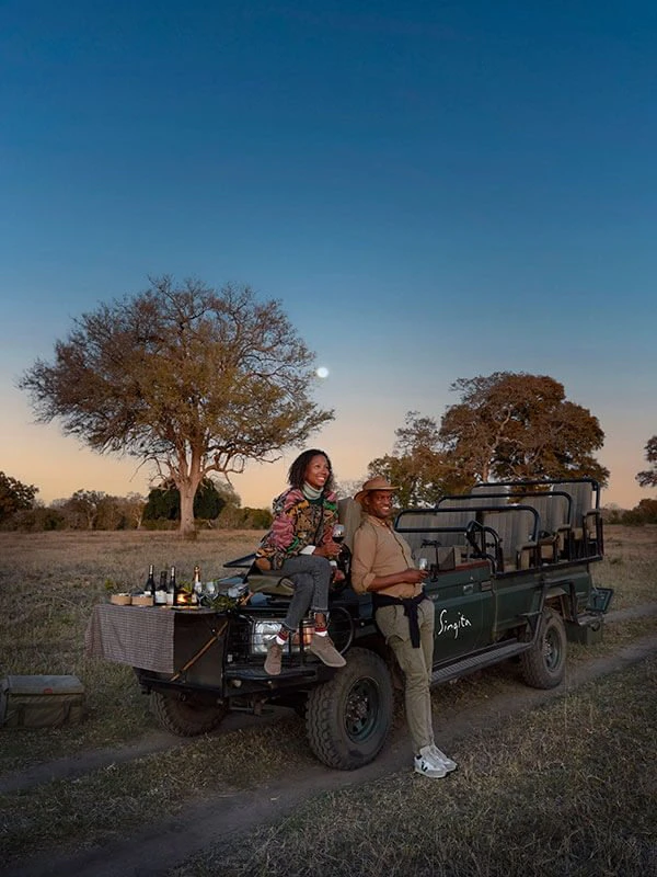 Couple enjoying a romantic sunset game drive with drinks setup on a luxury safari vehicle in the African wilderness.