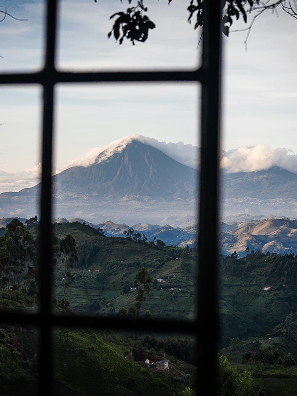 View of a mist-covered volcano and lush green hills through the window of a luxury safari lodge in Uganda, near Bwindi or Mgahinga National Park.