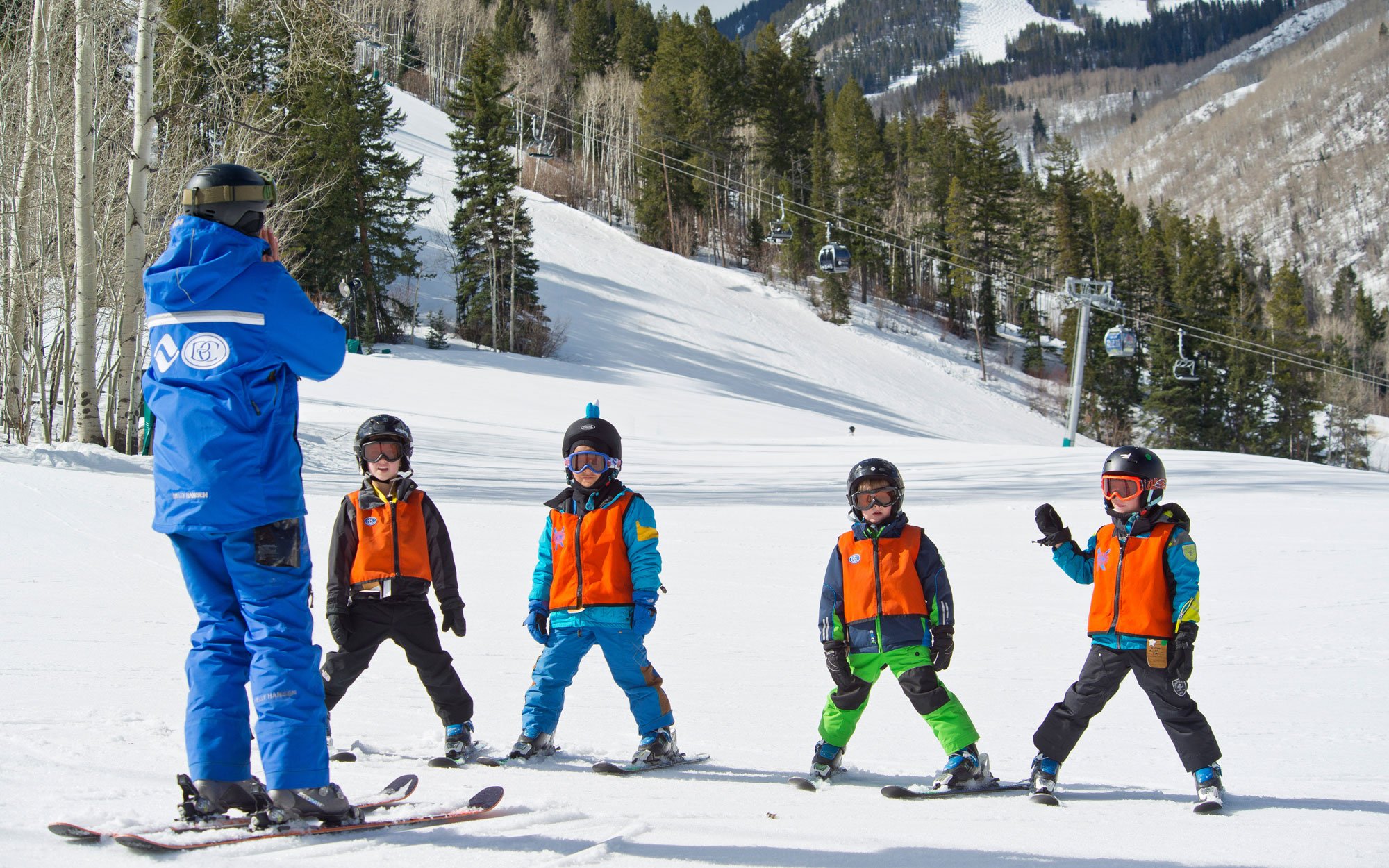 Beaver Creek ski school instructor in a blue jacket leading a beginner group lesson for four helmeted young children in orange ski-school bibs, all on skis on a groomed run with a chairlift and forested slopes behind